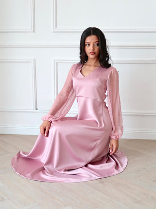 Full length satin and chiffon dress in soft pink with relaxed drape and sheer embellished sleeves, styled seated indoors